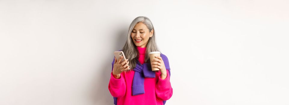 Trendy senior woman drinking coffee and using smartphone. Korean senior lady reading message on mobile phone and smiling, white background.