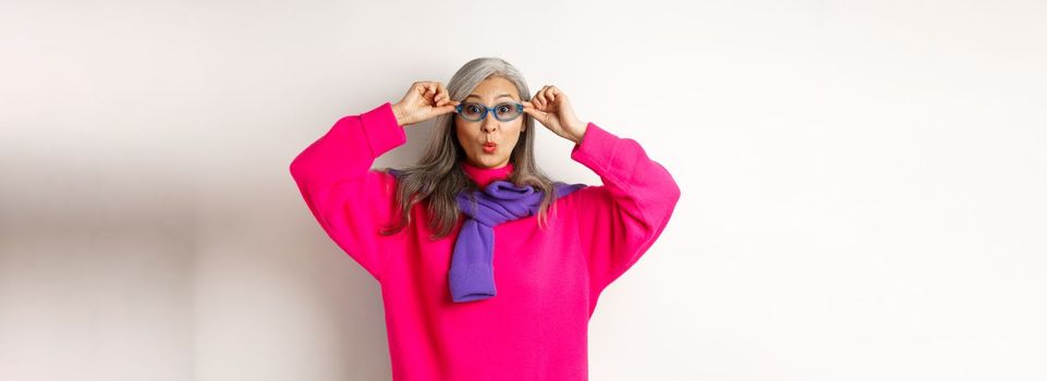 Trendy asian grandmother in sunglasses, pucker lips and looking surprised at camera, staring amazed at special offer, standing over white background.