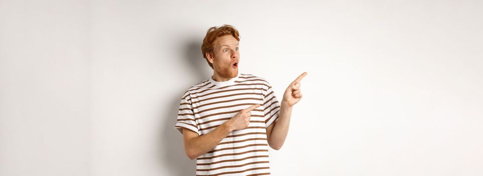 Check it out. Amazed redhead guy pointing and looking left at promotion banner, saying wow, standing over white background.