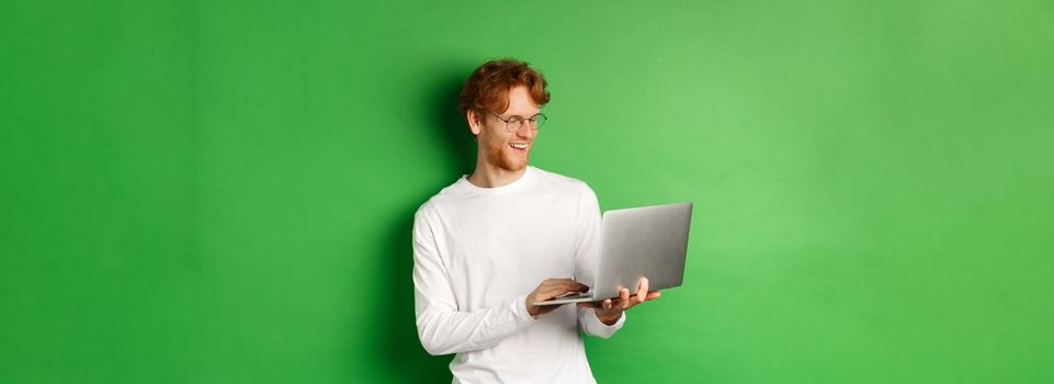 Smiling young man with red hair, wearing glasses, working on laptop and smiling, standing over green background.