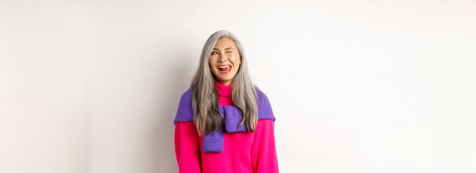 Funny and happy asian senior woman having fun, showing tongue and winking cheeky, looking left at logo, standing over white background.