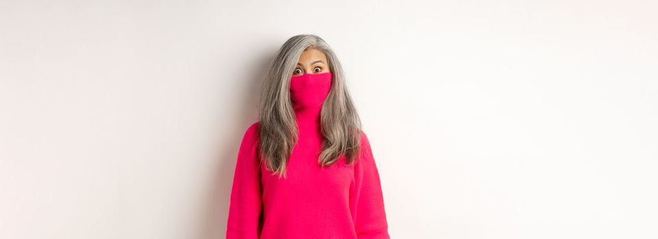 Funny and cute asian senior woman pull sweater collar on face and looking at camera, standing over white background.