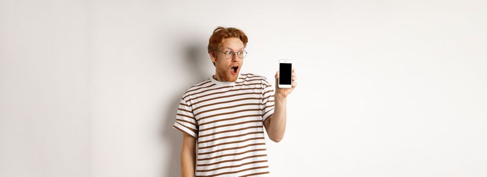 Technology and e-commerce concept. Surprised and shocked redhead guy checking out online promotion, showing blank smartphone screen and drop jaw, white background.