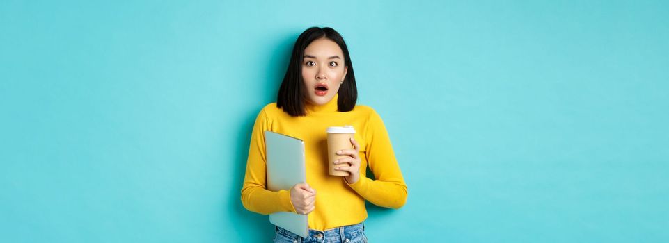 Shocked asian woman employee, drinking coffee from cafe takeaway, holding laptop, gasping and staring amazed at camera, standing over blue background.