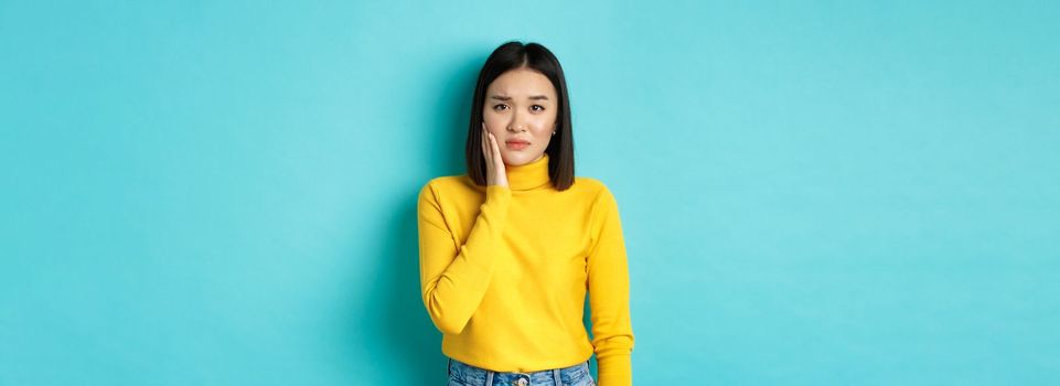 Portrait of asian young woman touching cheek and frowning, looking sad, being slapped in face, feeling painful toothache, standing over blue background.