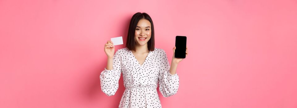 Cute asian woman shopping online, showing bank credit card and mobile screen, smiling and looking at camera, standing over pink background.