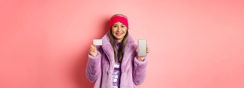 Online shopping and fashion concept. Fashionable asian senior woman showing black screen of smartphone and plastic credit card, smiling happy, pink background.