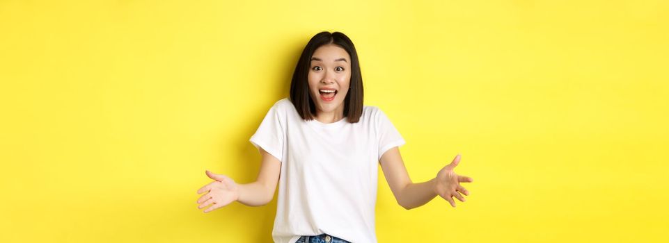 Excited young asian woman spread out hands as if holding large, big object, showing size and look amazed, standing over yellow background.