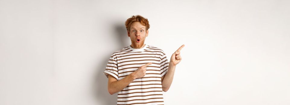 Amazed man with ginger hair checking out promo offfer, pointing left at logo and smiling at camera, standing over white background.