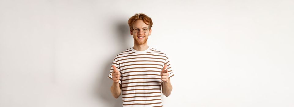 Young man with red hair and glasses pointing fingers at camera and smiling, need you, standing over white background.