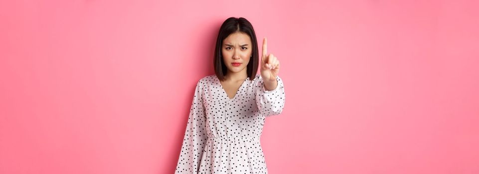 Serious and angry asian woman telling to stop, frowning and showing finger in disapproval, prohibit something, standing in dress against pink background.