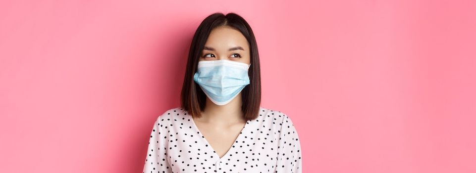 Covid-19, pandemic and lifestyle concept. Beautiful asian female model in medical mask laughing, smiling and looking left at copy space, standing over pink background.