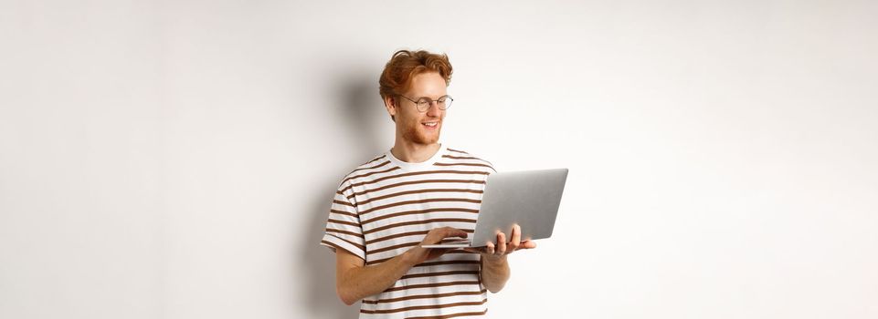 Young redhead freelancer working on laptop, typing on computer keyboard and smiling, standing over white background.