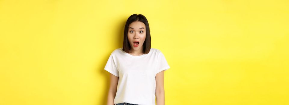 Beauty and fashion concept. Surprised asian girl open mouth and staring at camera, drop jaw impressed, standing over yellow background.