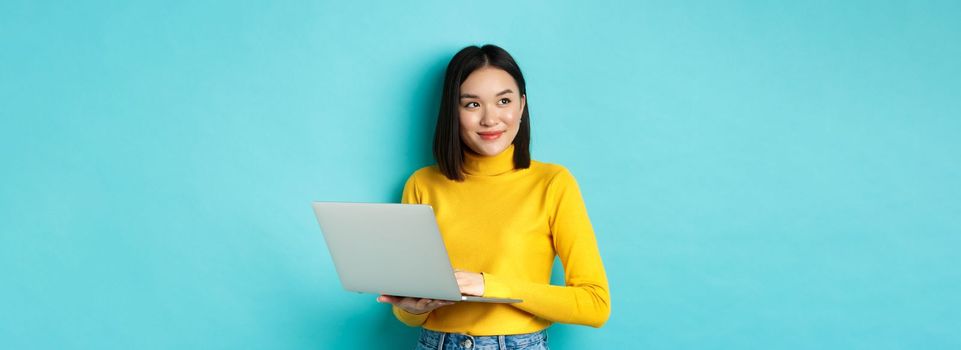 Image of stylish korean woman working on laptop and looking away with dreamy smile, standing with computer against blue background.