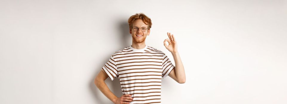 Confident smiling man with red hair assuring you, showing OK sign, guarantee quality, recommending something good, standing over white background.