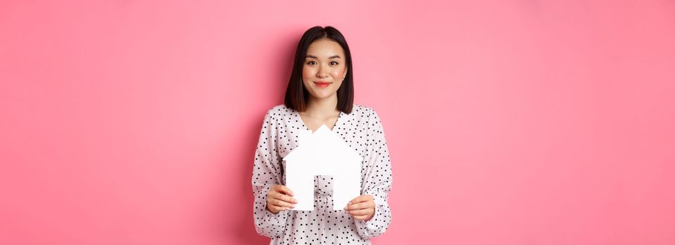 Real estate. Adult asian woman searching for home, holding house model and smiling, promo of broker company, standing over pink background.