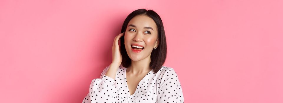Close-up of flirty asian woman sighing, looking dreamy left and smiling, standing over pink background. Copy space