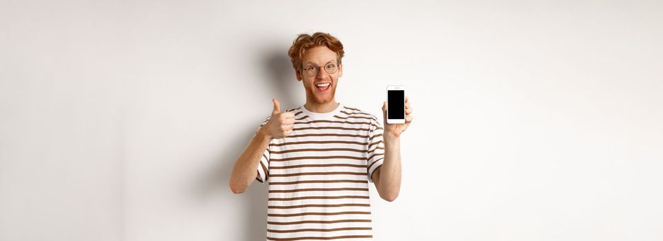 Technology and e-commerce concept. Young man with red hair showing thumbs-up and blank smartphone screen, recommending app, white background.