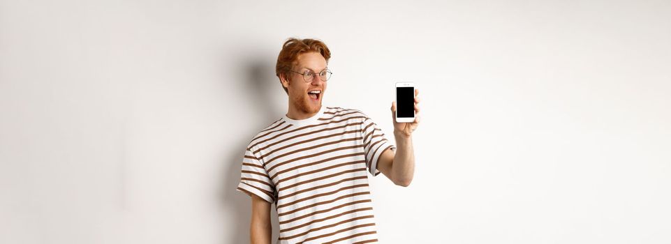 Technology and e-commerce concept. Happy young redhead man in glasses showing blank smartphone screen, looking amazed, standing over white background.