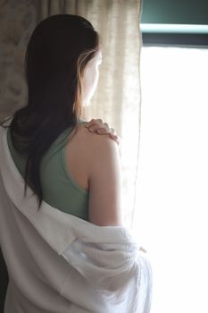 Beautiful woman in white silk pyjama standing near window in a cozy room. emotional portrait.