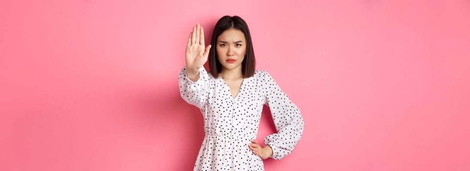 Angry asian woman tell stop, extend arm to prohibit or disapprove something, frowning displeased, standing over pink background.