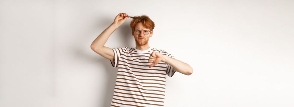 Young redhead man showing his messy haircut and thumbs-down, need hairdresser, standing over white background.