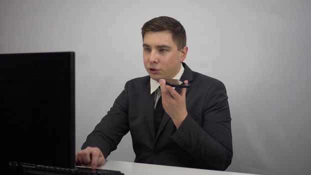 A young businessman records a voice message on a smartphone. A man in a suit sits in front of a computer and chats on his phone. 4k