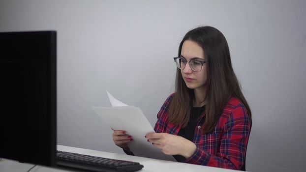 Office worker looks at documents at the workplace in the office. Young woman in glasses and a red shirt in front of a PC. 4k