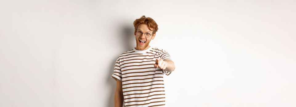 Image of funny redhead guy in glasses laughing at you, pointing finger camera and making fun of someone, standing over white background.