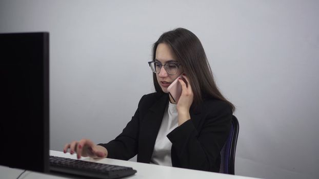 A young businesswoman works at a pc and speaks on the phone. A woman in a suit and glasses communicates on a mobile phone. 4k