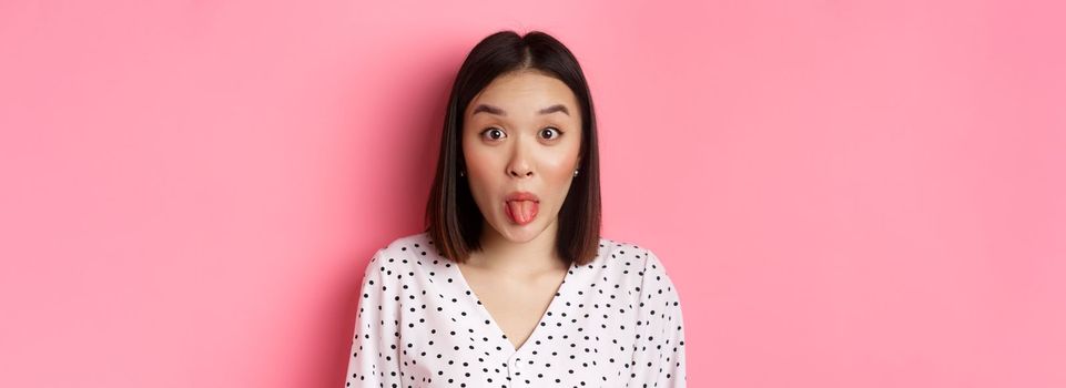 Beauty and lifestyle concept. Close-up of funny and cute asian woman showing tongue, staring at camera silly, standing over pink background.