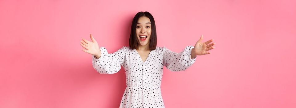 Happy asian girl stretching hands to camera, reaching for hug, smiling and wanting to hold something, standing over pink background in dress.