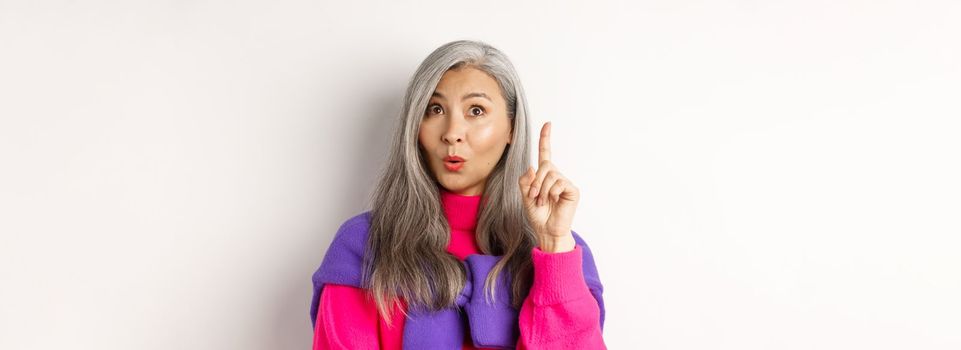 Close up of elderly asian woman in stylish hipster clothes, raising finger and looking up, having an idea, standing against white background.