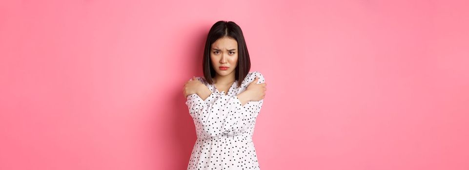 Timid and offended asian girl cross arms on chest, staring defensive and insulted at camera, standing in dress over pink background.