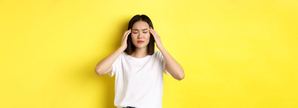 Portrait of asian woman close eyes and frown from migraine, touching head, feeling dizzy from headache, standing over yellow background.