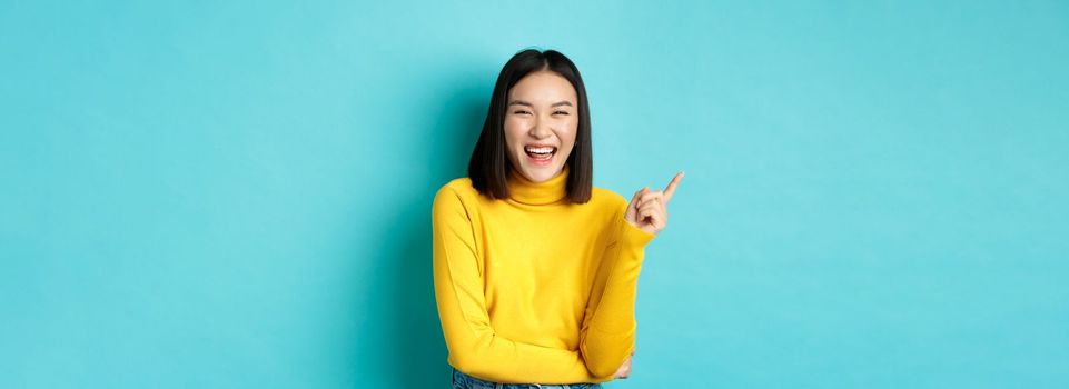 Image of carefree pretty korean girl laughing and looking happy, pointing finger at upper left corner promo, standing against blue background.