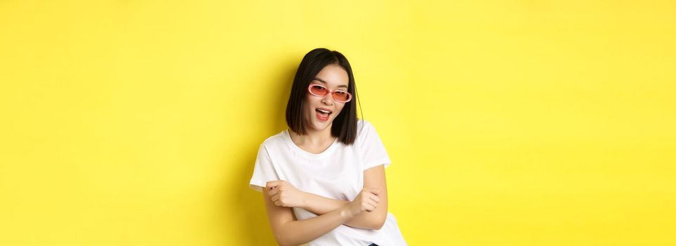 Fashion and lifestyle concept. Sassy and confident asian woman in trendy sunglasses looking self-assured at camera, standing over yellow background.