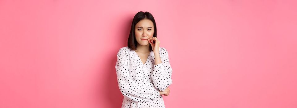 Brunette asian woman in dress looking displeased, frowning and zipping mouth, seal lips with promise, standing over pink background.