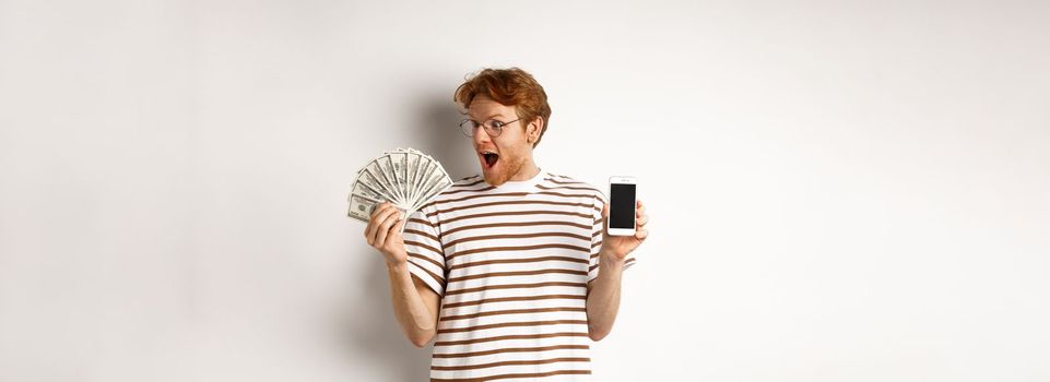 Amazed redhead man showing smartphone app on blank screen and money, winning prize cash online, standing over white background.