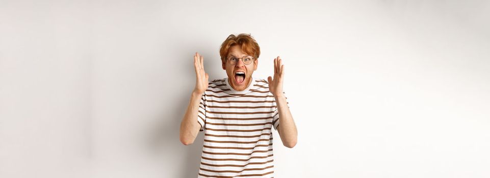 Angry young man with red hair shouting at camera, screaming and looking outraged, shaking hands, standing over white background.