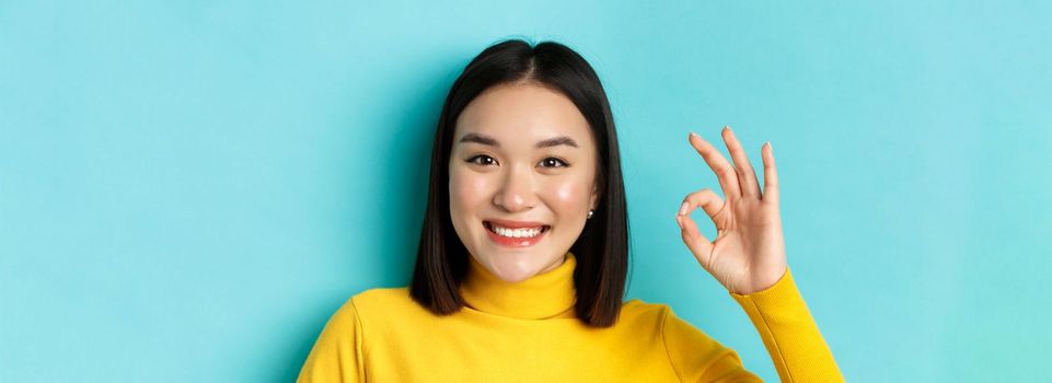 Beauty and makeup concept. Close up of happy asian woman with healthy shiny skin showing OK sign and smiling satisfied, standing over blue background.