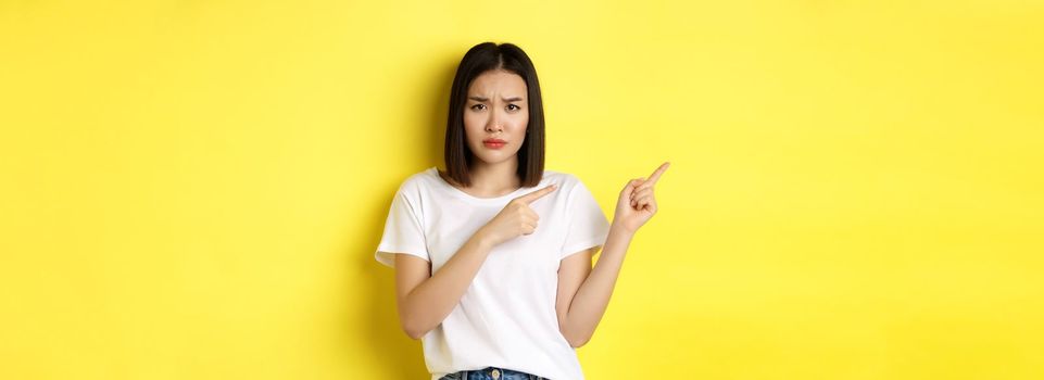 Beauty and fashion concept. Beautiful asian woman in white t-shirt pointing fingers left, standing over yellow background.