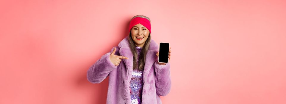 Online shopping and fashion concept. Stylish asian mature lady showing blank smartphone screen, pointing at phone and smiling, pink background.