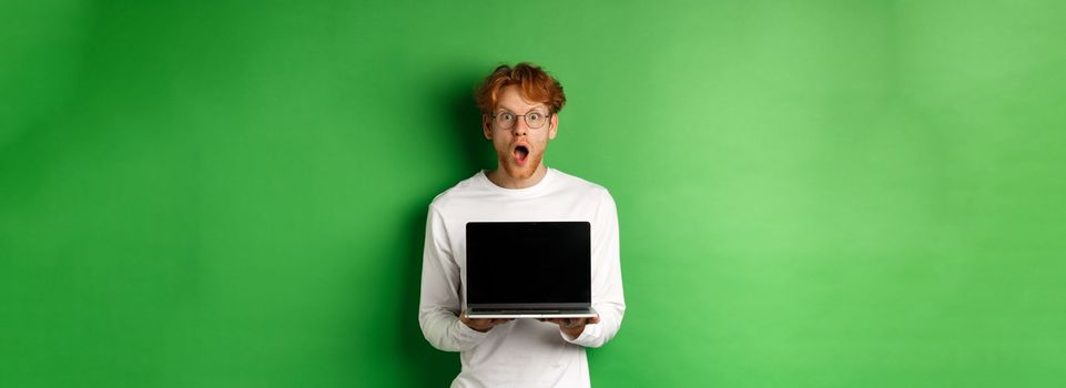 Surprised redhead man in glasses showing laptop blank screen, showing online promo and drop jaw, staring at camera amazed, green background.