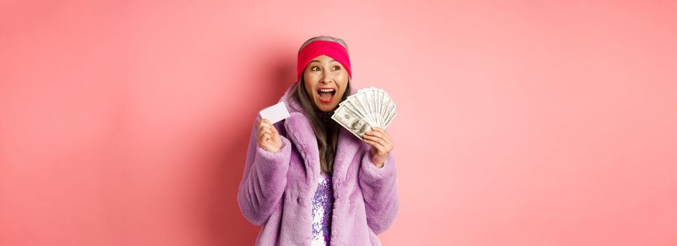 Shopping and fashion concept. Asian senior woman scream happy like winner, holding dollars money and plastic credit card, looking excited, pink background.