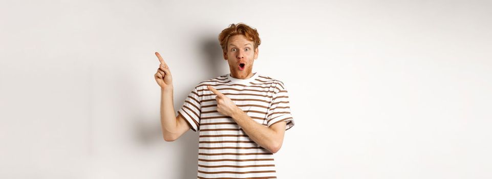 Excited man with curly red hair showing promo offer and looking amazed, pointing fingers at upper right corner logo, white background.