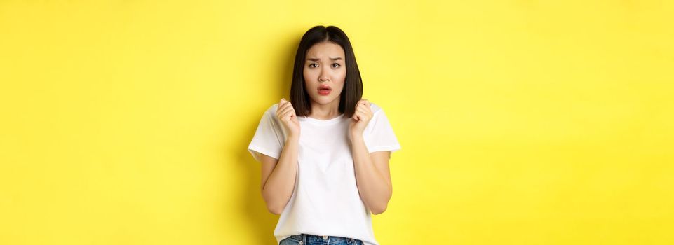 Beauty and fashion concept. Scared timid asian girl looking anxious, gasping startled and standing over yellow background.