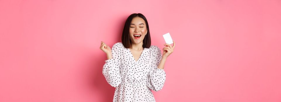 Shopping concept. Excited asian woman feeling satisfaction, holding credit card and rejoicing, standing over pink background.