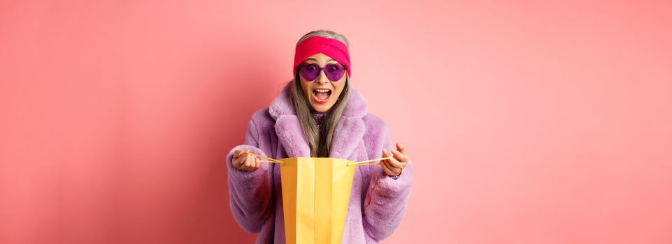 Shopping and fashion concept. Stylish asian elderly woman in sunglasses and faux fur coat open paper bag with gifts, looking amazed at camera, pink background.